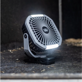 A small portable fan with integrated LED light bars, shown lit and standing on a dark textured surface. The fan has a white outer housing, black inner grille, and a circular black center logo featuring a stylized 'W'.
