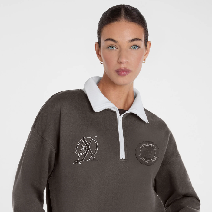 A woman wearing a dark olive gray quarter-zip sweatshirt with a white collar. The sweatshirt features a white embroidered logo on the left chest and a circular patch on the right chest.