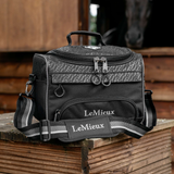 LeMieux AW26 Elite ProKit Lite Grooming Bag Varient Black. A black LeMieux equestrian grooming bag with a patterned top panel and a prominent adjustable shoulder strap, resting on a wooden stable bench with a horse stable in the background.