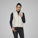 A woman modeling a light cream quilted puffer vest over a black long-sleeve top and black riding breeches, photographed against a solid light gray studio background.