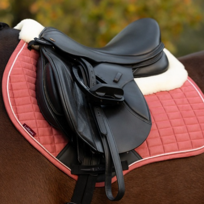 LeMieux AW26 Merino+ Suede Classic Close Contact Square Varient Spiced Earth. A black leather equestrian saddle resting on a pink quilted horse pad with white fleece lining, shown on a horse against a blurred outdoor background.