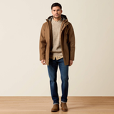 A man wearing a brown hooded jacket over a beige sweater, blue jeans, and brown boots, standing against a plain beige studio background with a light wood floor.