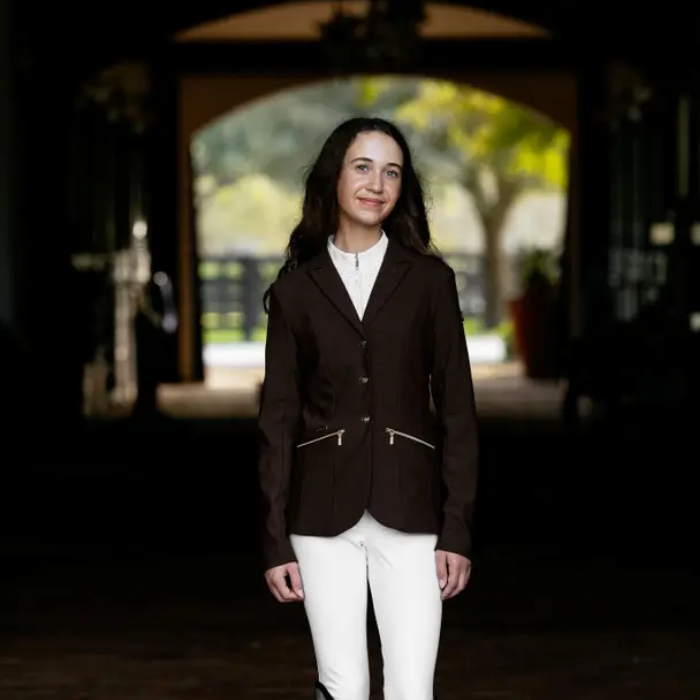 Woman wearing a dark brown equestrian jacket and white pants standing in a dimly lit area with an archway in the background.