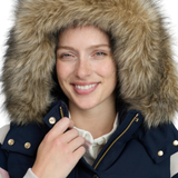 A smiling woman wearing a navy winter gilet with a large brown faux-fur hood and gold-tone snaps and zippers, photographed against a clean white background.