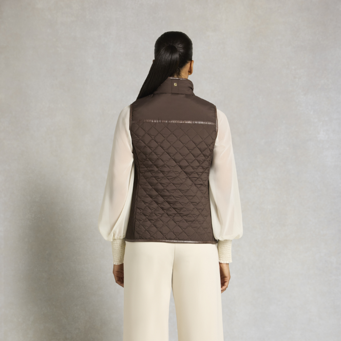 Holland Cooper Boughton Diamond Quilt Gilet Variant Colour Walnut.
A model shown from the back wearing a dark brown quilted puffer vest over a cream-colored long-sleeve blouse and matching cream trousers, set against a textured light gray studio background.