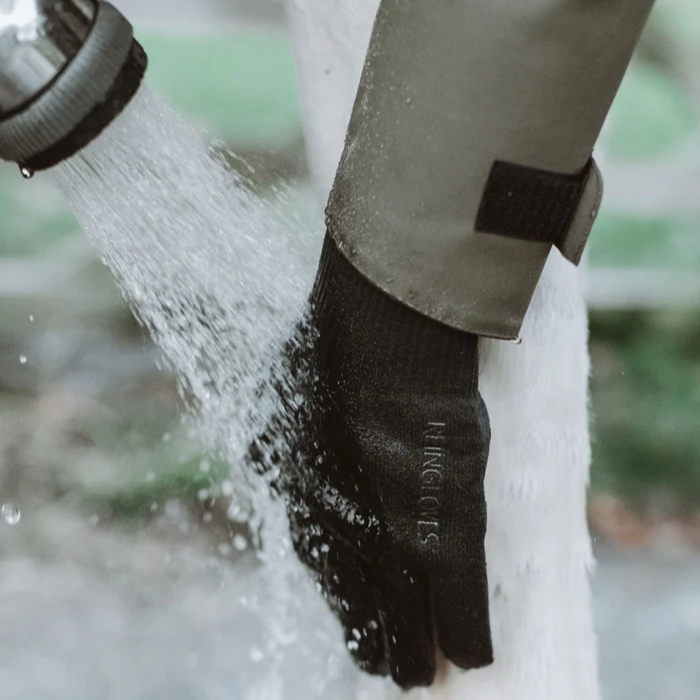A close-up of a black knit glove with a green waterproof sleeve being washed by running water from a hose. The brand name 'REINCOAT' is printed on the back of the glove.