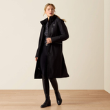 A woman models a long black hooded coat over a patterned top, black pants, and black knee-high boots in a minimalist studio setting with a beige wall and light wood floor.