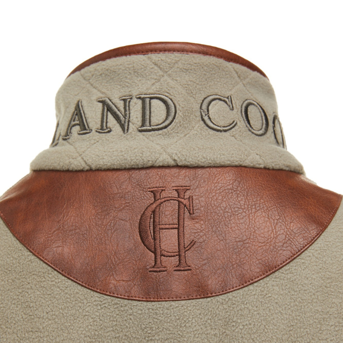 Holland Cooper Country Fleece Gilet Variant Sage. Close-up of the back of a jacket showing a sage fleece collar with dark green embroidered lettering, a brown leather band, and an embossed monogram logo on a white background.