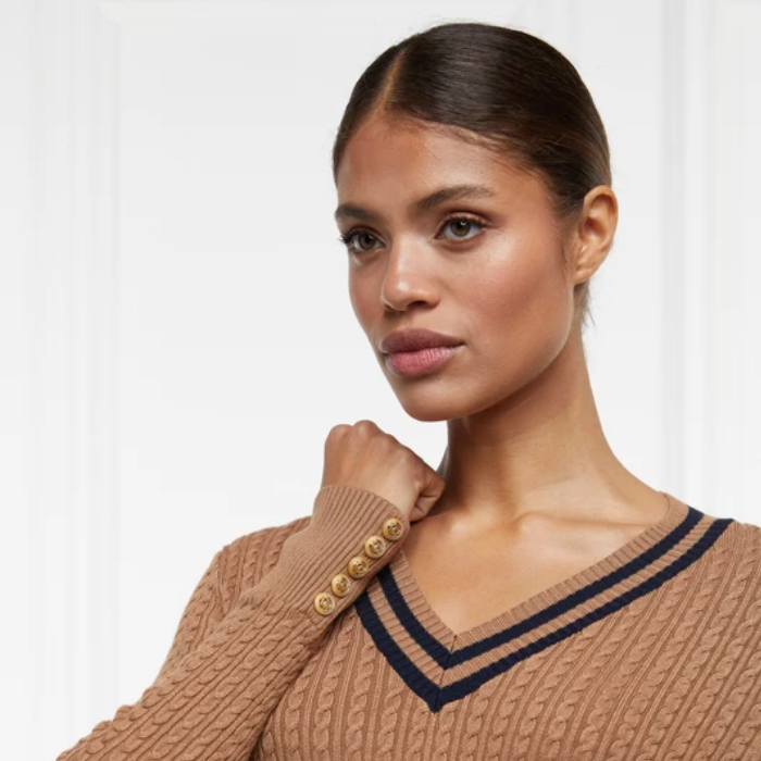 Holland Cooper Zoe Knit Jumper Variant Dark Camel. A woman wearing a tan cable-knit V-neck sweater with dark navy trim and decorative gold buttons on the sleeve, photographed against a clean white background.