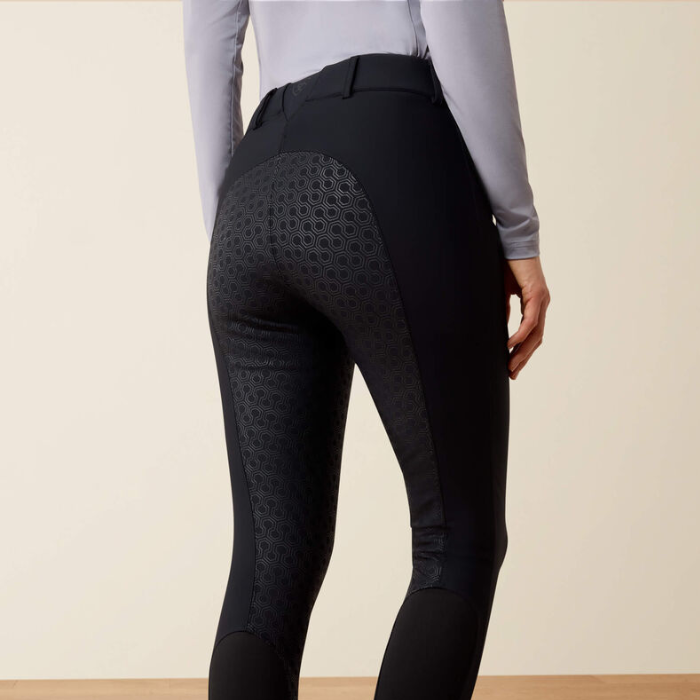 Rear view of a person wearing black equestrian riding breeches with textured patterned side panels and a light gray long-sleeve top, against a plain beige studio background.