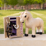 LeMieux AW26 Toy Pony Tack Locker Wooden Varient Brown. Small wooden tack box open with toy pony bridle hooked on the door with beige toy pony stood on a concrete floor with grass garden behind. 