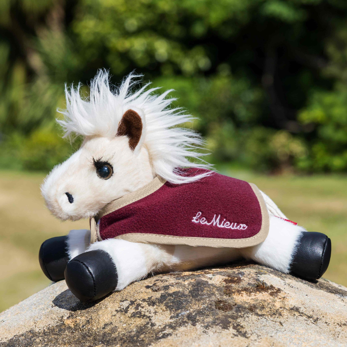 A plush toy horse wearing a burgundy and beige saddle blanket with the embroidered brand name 'Le Mieux', resting on a rock with a blurred green outdoor background.