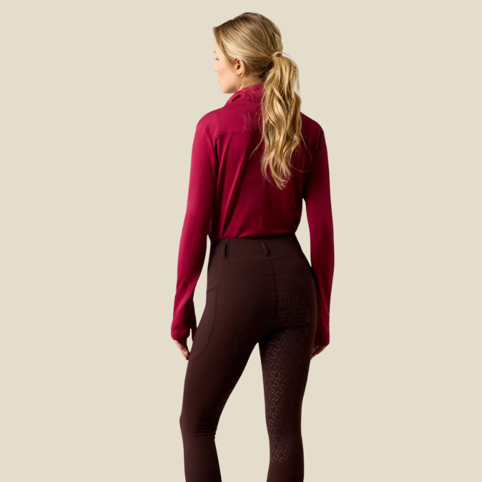 Ariat Women's Venture 2.0 Thermal Full Seat Tight Variant Chocolate Plum. A woman shown from the back wearing a dark burgundy long-sleeve athletic top and dark brown equestrian riding breeches with textured knee panels, set against a solid light beige background.