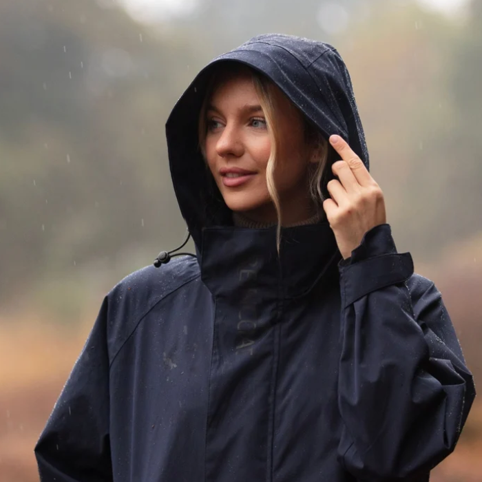 A woman wearing a dark navy hooded rain jacket stands outdoors in the rain, holding the edge of her hood with one hand.