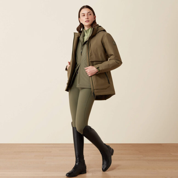 Lady wearing a khaki coloured jacket with a dark green zip against a beige background. 