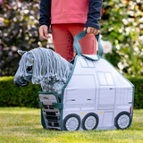 LeMieux AW26 Toy Pony Carry Bag Varient Printed Grey. A child wearing coral pink leggings and a pink top holds a LeMieux horse in a white and teal toy horse trailer on a green grass lawn with a blurred garden background.