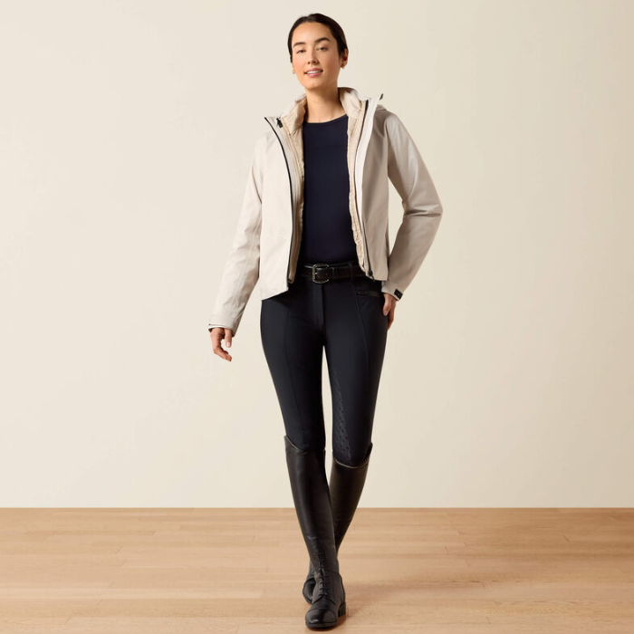 A woman modeling a light beige hooded jacket over a navy top and black riding pants and boots, standing in a studio with a warm off-white wall and light wood floor.