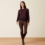 A woman modeling a dark plum long-sleeve knit sweater and matching dark brown riding tights with knee patches, paired with dark brown riding boots, against a warm beige studio background.