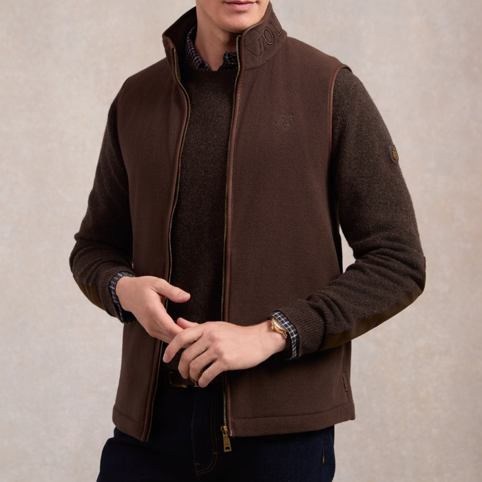 Holland Cooper Country Fleece Gilet (Men's) Variant Chocolate. Man wearing a brown fleece with Holland Cooper branding roun the collar, over a brown jumper on a grey background. 