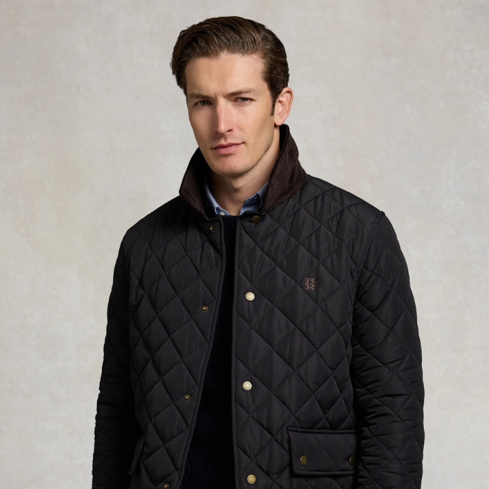 A man wearing a navy diamond-quilted jacket with a dark brown ribbed collar, gold-tone snap buttons, and a small embroidered logo on the chest, set against a textured light gray background.