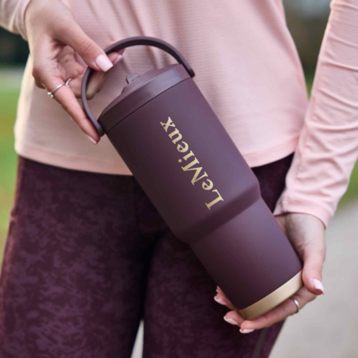 LeMieux AW26 Sports Flip Cup Varient Blackberry. A person holds a LeMieux travel tumbler with a dark plum body, gold lettering, and a gold base ring. The person is wearing a light pink long-sleeve top and dark plum leggings, with a blurred outdoor background.