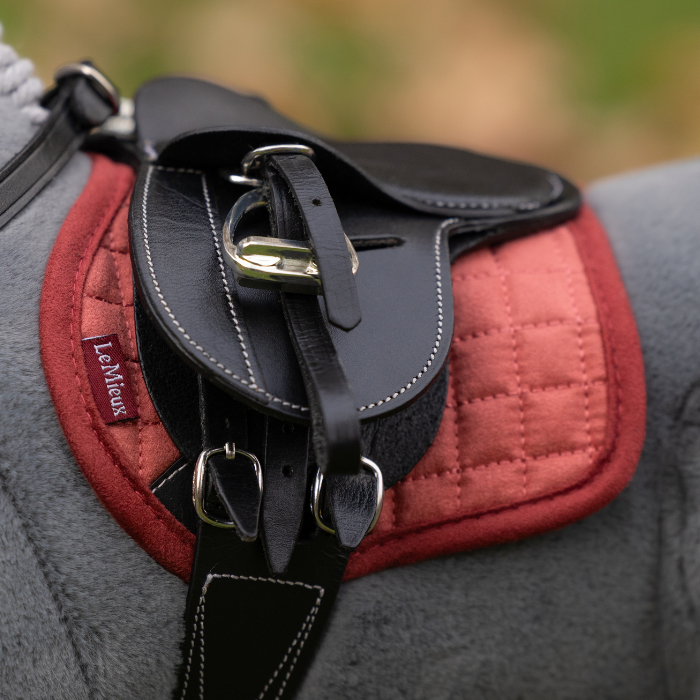 LeMieux AW26 Toy Pony Pad Variant Colour Spiced Earth.
A close-up of a toy pony black leather equestrian saddle pad with white stitching and silver buckles, layered over a red quilted saddle pad on a gray plush toy pony, with a blurred outdoor background.