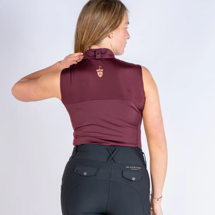 Woman wearing a maroon sleeveless top and dark gray riding pants on a white background