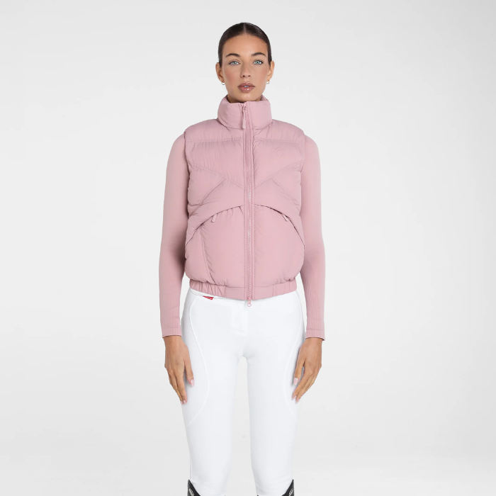A model wearing a dusty pink cropped quilted puffer vest over a matching pink long-sleeve top, paired with white riding breeches, against a plain white studio background.