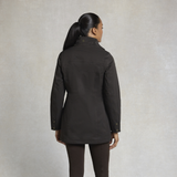 Holland Cooper Bramwell Waterproof Country Coat Variant Colour Bournville.
A woman shown from the back wearing a dark brown mid-length jacket and dark brown leggings, standing against a textured light gray studio background.