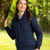 LeMieux AW26 Ladies Team Jacket Variant Colour Navy. A model wearing navy jacket leaning against a tree on faded garden background. 