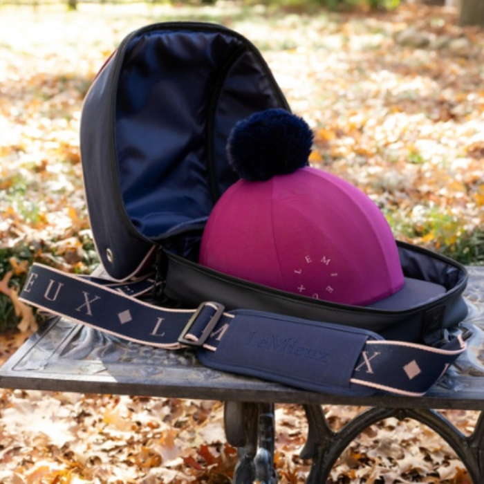LeMieux AW26 Hat Bag Varient Blackberry. A pink LeMieux riding cap with a dark navy blue pom-pom sits inside an open navy blue equestrian saddlebag. The bag features a wide, adjustable navy blue shoulder strap with 'LE MIEUX' branding printed in white. 