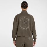 A model viewed from the back wearing a brown cropped zip-up jacket and matching sweatpants. The jacket features a large circular graphic on the back reading 'AZTEC DIAMOND SPORTING GOODS' with a stylized 'AD' logo in the center.