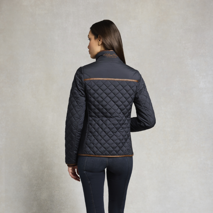 Holland Cooper Boughton Diamond Quilt Jacket Variant Colour Ink Navy.
A woman shown from the back wearing a dark navy quilted jacket with tan contrast stitching, paired with dark navy pants, against a textured light gray studio background.
