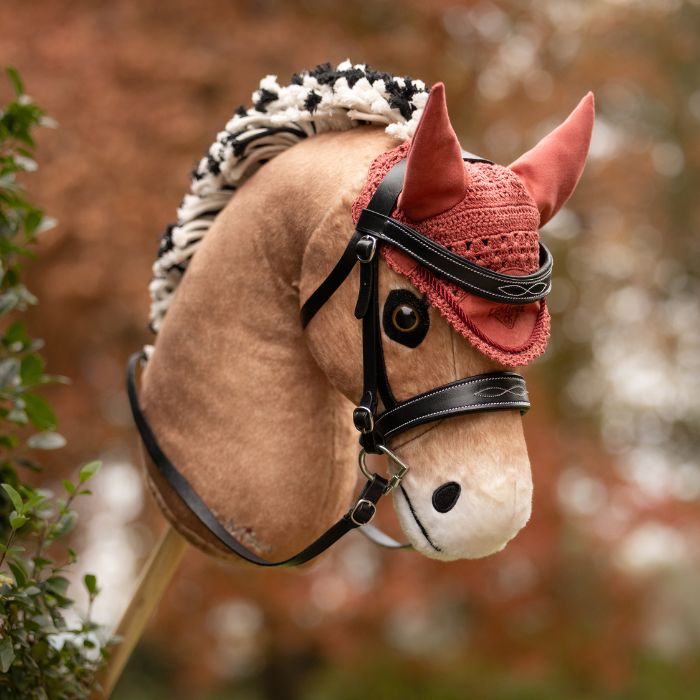 LeMieux AW26 Hobby Horse Fly Hood Variant Colour Spiced Earth. A plush toy horse head on a stick, light brown colour with wool short briaded mane and a spiced earth colour coloiur fly hood and bridle on a faded garden background.