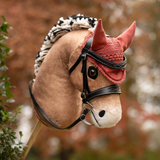 LeMieux AW26 Hobby Horse Fly Hood Variant Colour Spiced Earth. A plush toy horse head on a stick, light brown colour with wool short briaded mane and a spiced earth colour coloiur fly hood and bridle on a faded garden background.