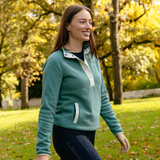 LeMieux AW26 Piper Popper Fleece Varient Aloe. A woman wearing a sage green quarter-zip fleece pullover with white contrast stitching and a white zipper pull, walking in a sunny park with autumn trees in the background. LeMieux branding is visible on her leggings.