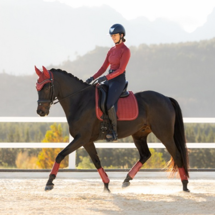 LeMieux AW26 Suede Dressage Square Varient Russet. A rider in equestrian apparel sits on a dark horse in an outdoor riding arena. The horse wears red protective leg wraps and a red saddle pad, with a matching red horse veil.