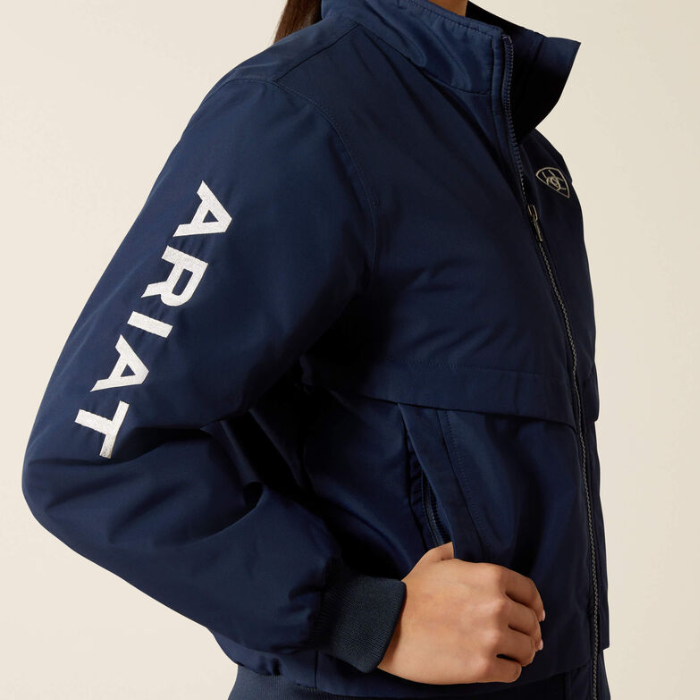 A close-up of a person wearing a navy blue jacket with large white vertical ARIAT branding on the left sleeve and a small circular logo on the left chest, against a plain light beige background.