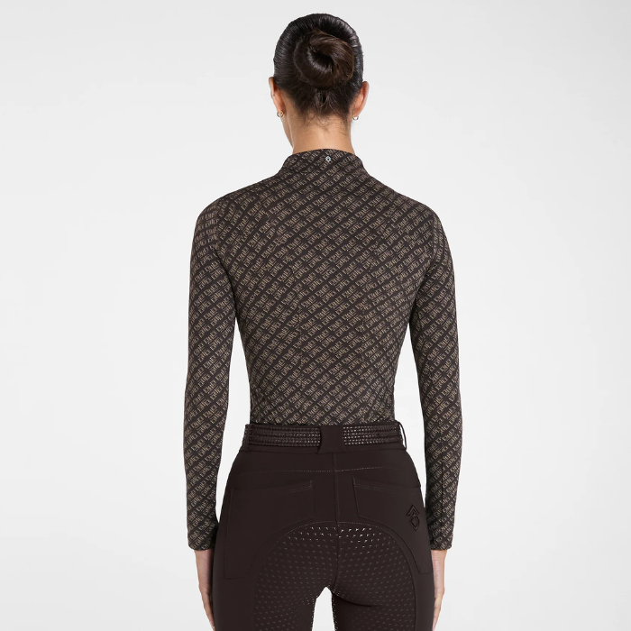 Rear view of a woman wearing a long-sleeve equestrian riding shirt with a dark brown and black repeating monogram pattern, paired with dark riding breeches, against a light gray studio background.