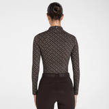 Rear view of a woman wearing a long-sleeve equestrian riding shirt with a dark brown and black repeating monogram pattern, paired with dark riding breeches, against a light gray studio background.