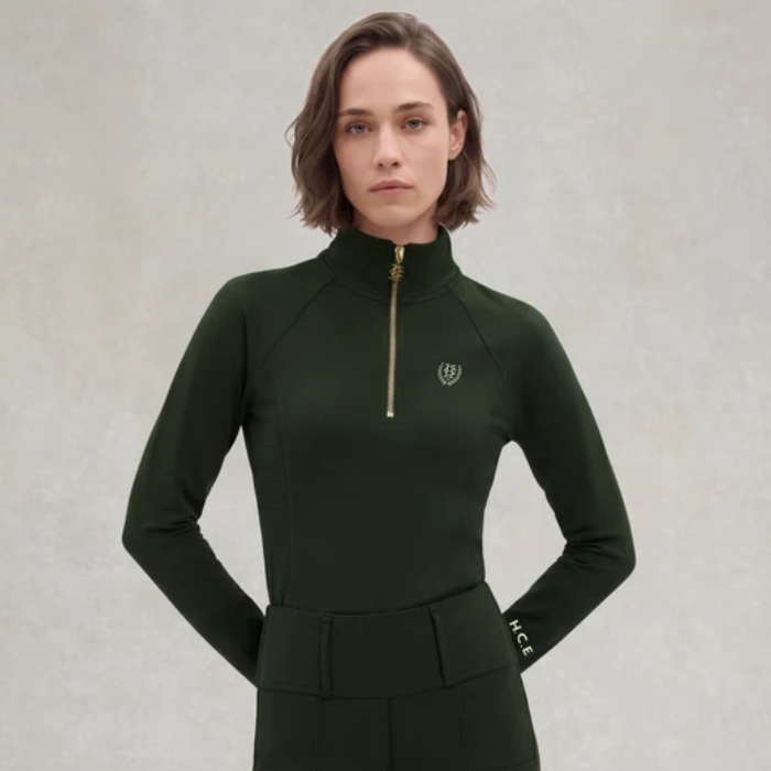 Holland Cooper Cranborne Long Sleeve Base Layer Variant Dark Olive. A woman modeling a dark forest green long-sleeve athletic top with a half-zip high collar, a small gold chest logo, and 'HCE' printed in white on the cuff, set against a textured light gray background.