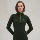 Holland Cooper Cranborne Long Sleeve Base Layer Variant Dark Olive. A woman modeling a dark forest green long-sleeve athletic top with a half-zip high collar, a small gold chest logo, and 'HCE' printed in white on the cuff, set against a textured light gray background.