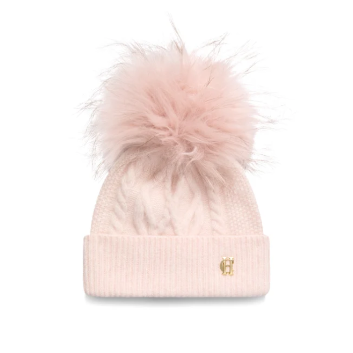 Holland Cooper Sloane Cable Bobble Hat variant Soft Pink. A light pink cable-knit beanie with a large fluffy faux-fur pom-pom on top and a small gold monogram logo on the front cuff, shown against a plain white background.