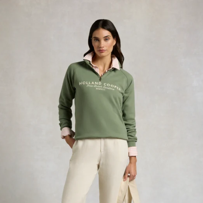A woman models a green quarter-zip sweatshirt with a pink inner collar and white 'HOLLAND COOPS' text on the chest, paired with cream pants against a textured gray studio background.