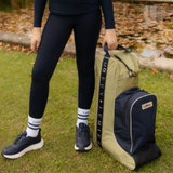 LeMieux AW26 Boot & Hat Bag Varient Gooseberry. A person outdoors on grass beside a pond holds a large boot and hat bag with a olive green back section and navy front section. 'LeMieux' is displayed down the side. 