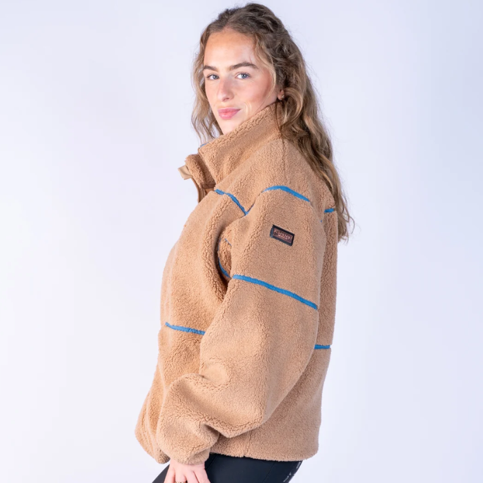 Blackfort AW26 Layla Fleece Variant Tan & Glacier. Girl wearing a an zip up fleece with Blue stripe detailing and a small rose gold logo on the sleeve, agaisnt a grey background. 