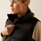 Ariat Women's Ultrapuff Down Vest Variant Black. A close-up of a woman wearing a black puffer vest over a beige long-sleeve top, shown from the chest up against a plain light beige background.