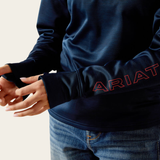 Ariat Youth Team 1/2 Zip Pullover Variant Navy. A close-up of a person wearing a navy blue long-sleeve athletic top with 'ARIAT' outlined in red on the left sleeve, paired with blue jeans against a light beige background.