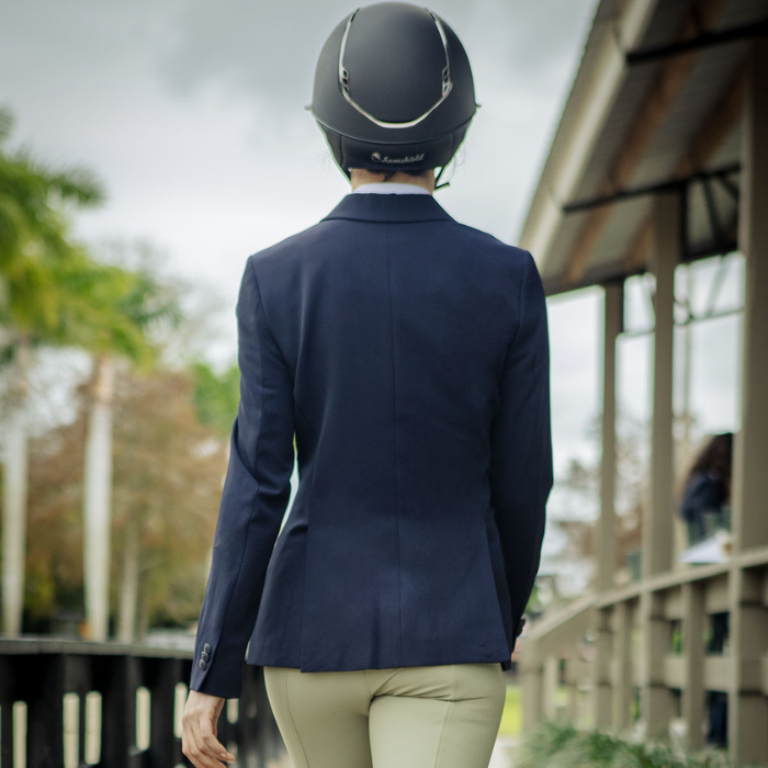 Person wearing a navy blue equestrian jacket and helmet, standing on a path with greenery and a building in the background.