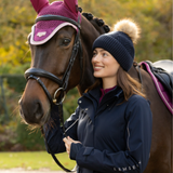LeMieux AW26 Stormwear Performance Riding Coat Varient Navy. A woman in a navy blue knit beanie with a pom-pom and a navy blue riding jacket stands next to a brown horse wearing a purple fly veil and bridle, outdoors in a park-like setting.